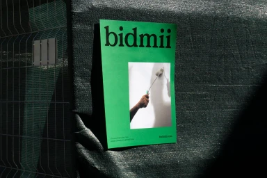 Green Bidmii poster featuring a hand painting a white wall, affixed to a dark green mesh construction fence.