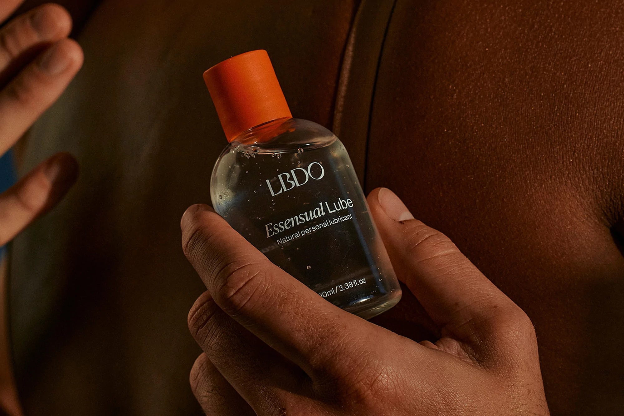 Close-up of a clear bottle of LBDO lubricant with an orange cap, held by a person with tan skin and a dark background.