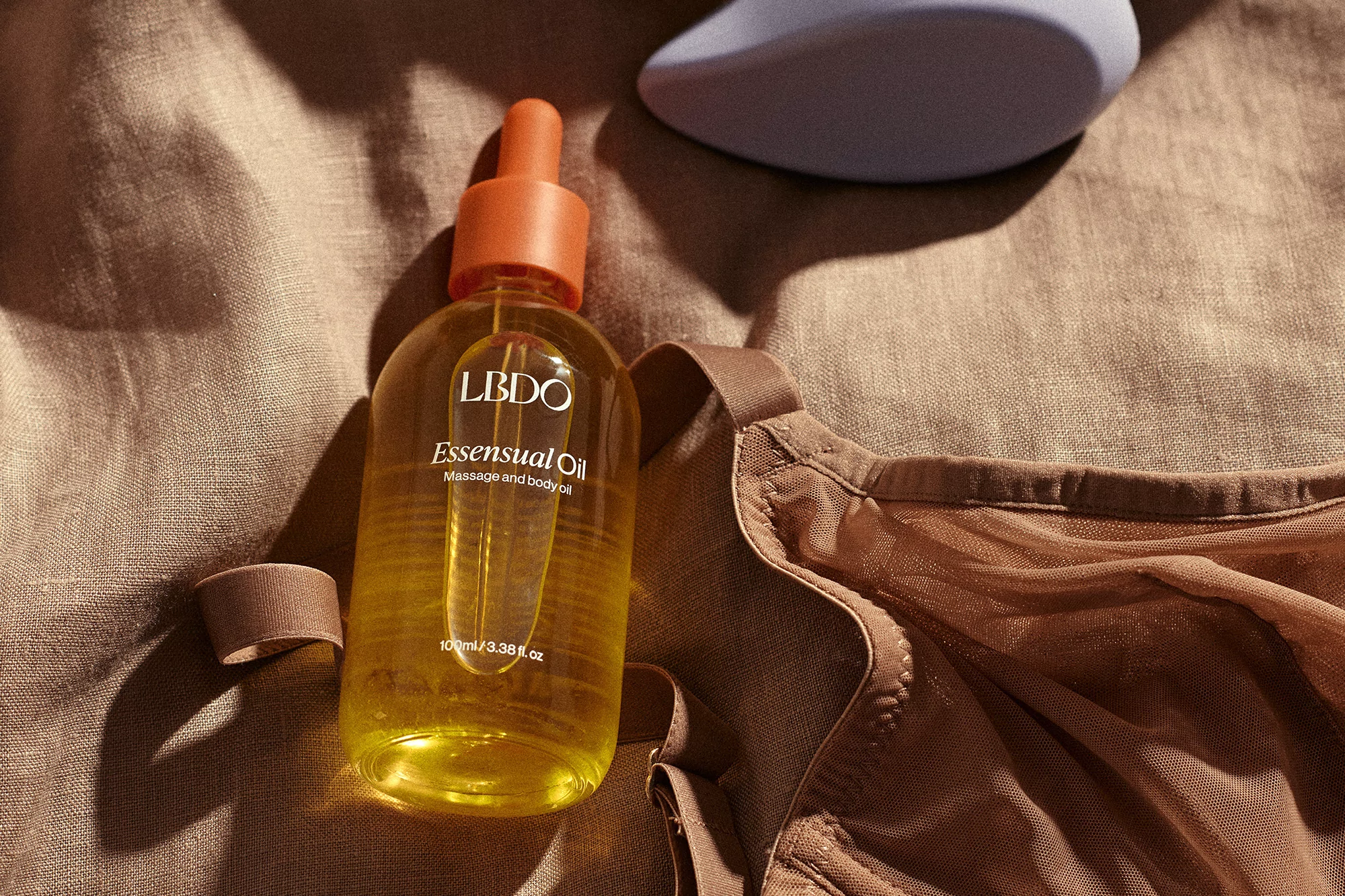 Close-up studio shot of LBDO essential oil bottle with an orange dropper, beige bra, and violet pleasure object on a linen surface.