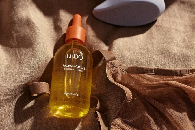 Close-up studio shot of LBDO essential oil bottle with an orange dropper, beige bra, and violet pleasure object on a linen surface.