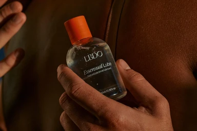 Close-up of hands holding a clear personal lubricant bottle with an orange cap against a warm-toned body.