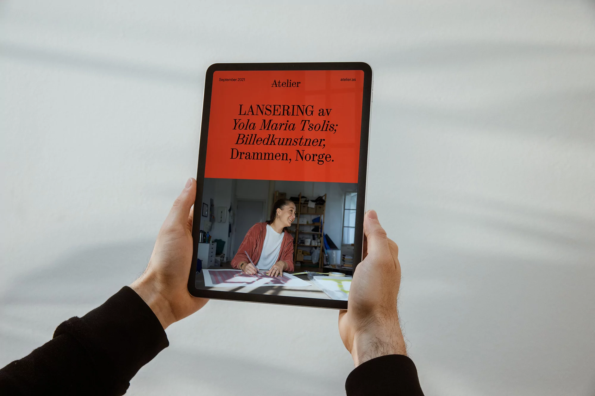 A tablet displays an Atelier magazine cover featuring an artist; it's held by two hands against a white backdrop, creating a modern, informative scene.