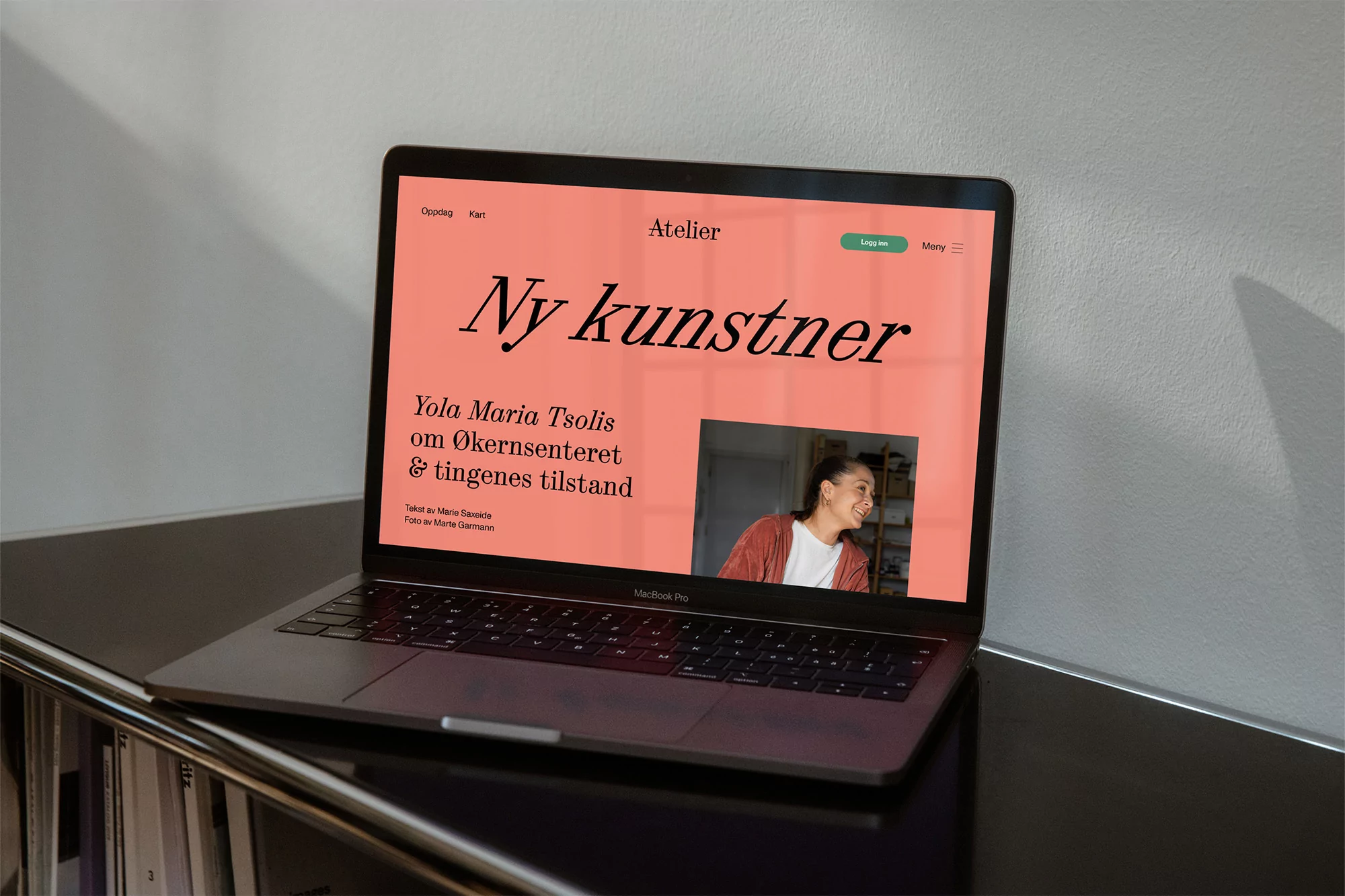 Laptop displaying 'Atelier' website with the headline 'Ny kunstner' in front of a white wall and a shelving unit, showcasing digital design.