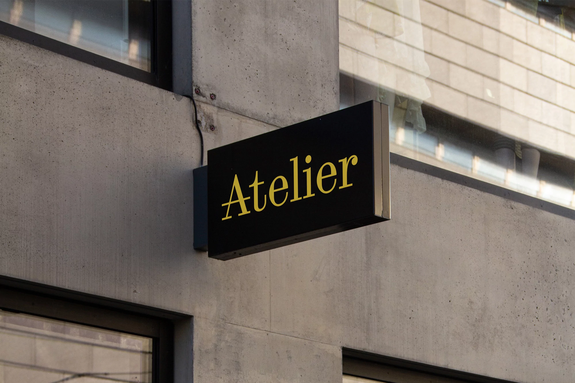 Black and gold 'Atelier' sign mounted on the grey concrete exterior wall of a building with windows in the background.
