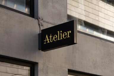 Black rectangular sign with yellow text Atelier mounted on a raw concrete exterior wall next to glass windows