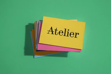 A stack of colorful paper cards featuring the word Atelier on top, resting on a green surface with visible shadows.