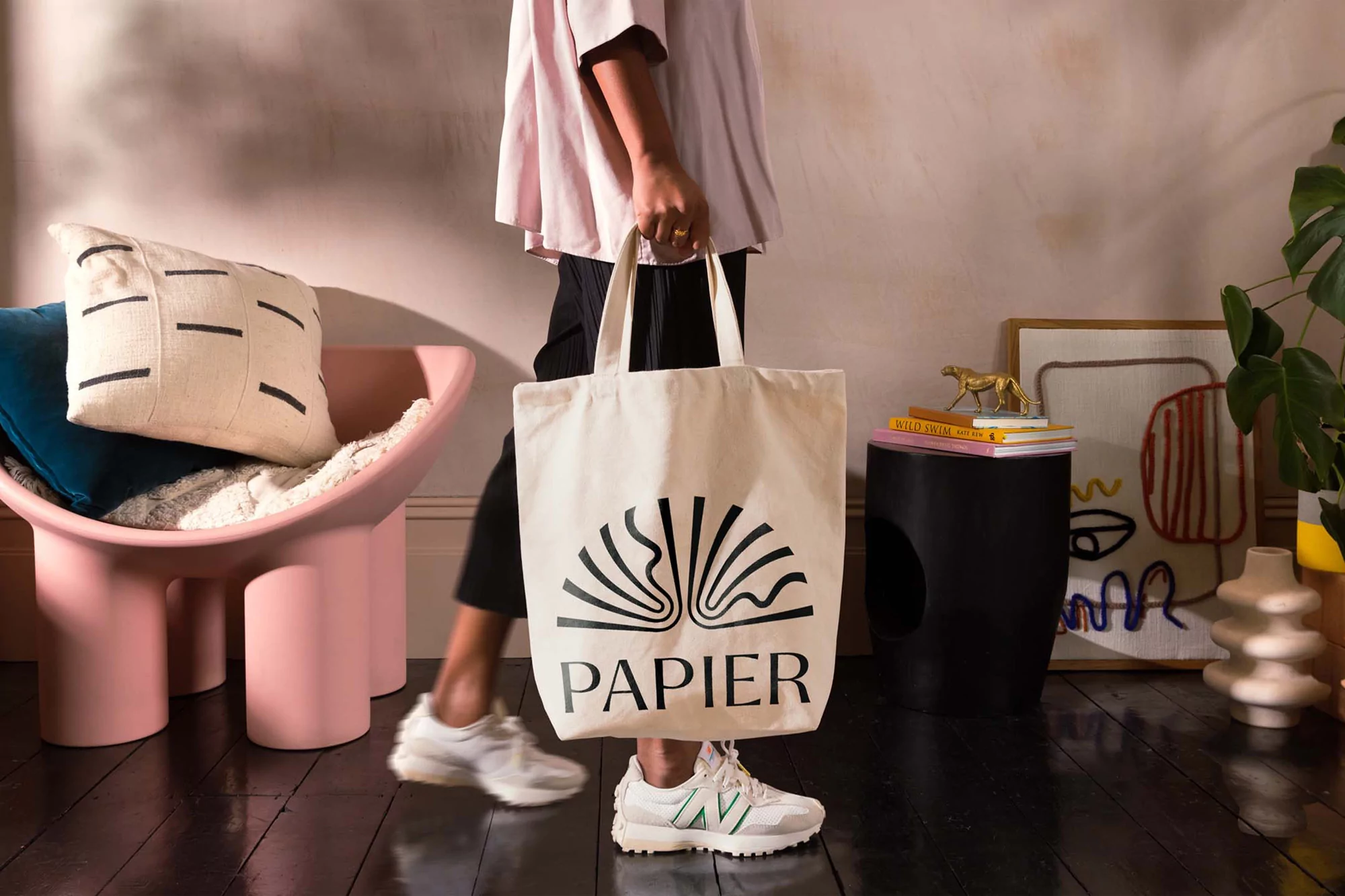 Person carrying a 'Papier' branded tote bag in a stylish interior setting, with a pink chair, patterned pillows, and modern decor.