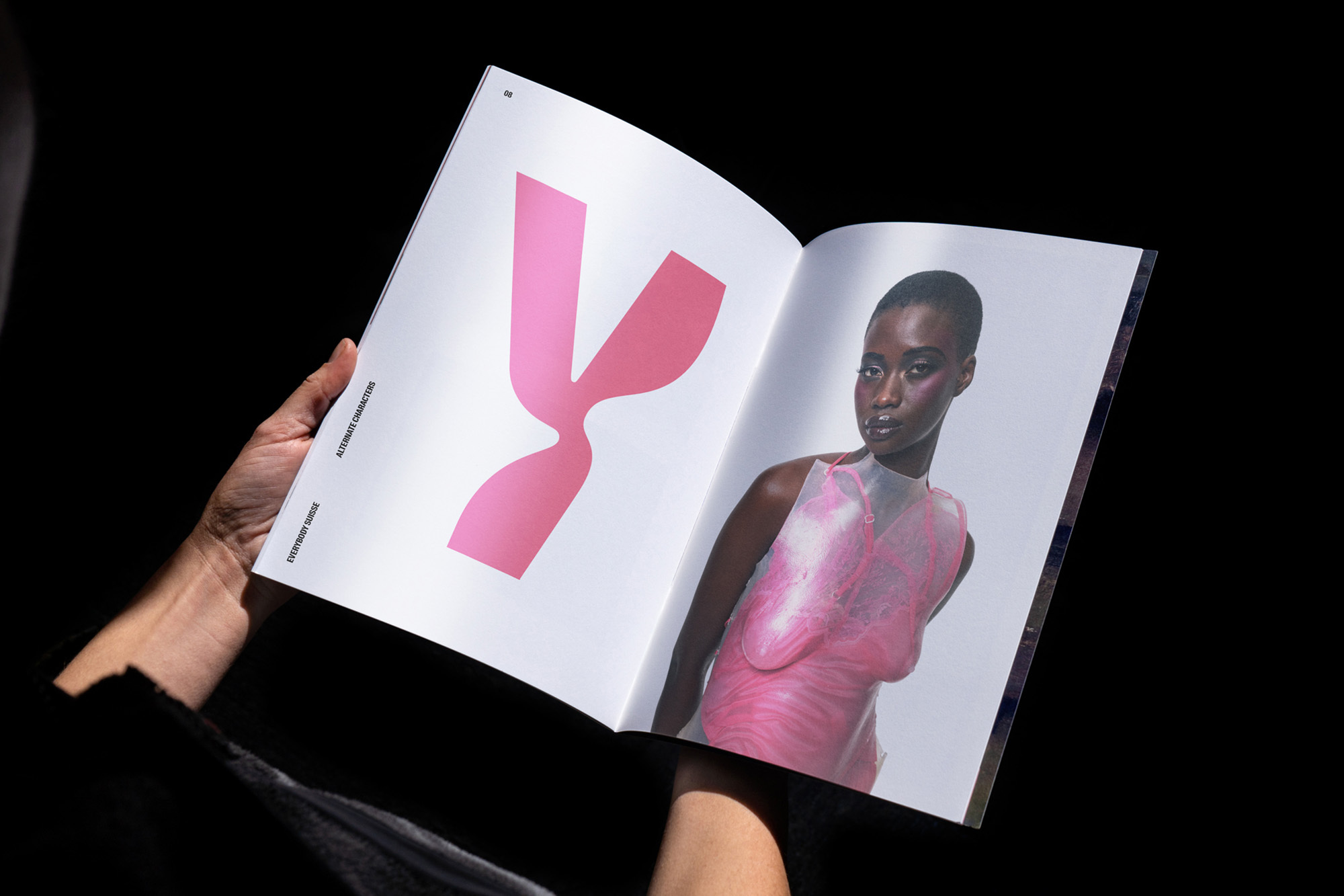 Open magazine with an image of a woman in a pink top and a pink graphic, held by a person against a dark background.