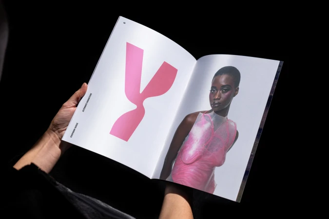 Open magazine with an image of a woman in a pink top and a pink graphic, held by a person against a dark background.