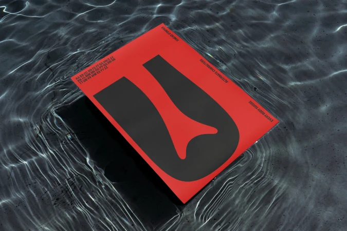 A red album cover featuring a black 'U' lies on a surface with a water texture, displaying refractions and highlights.