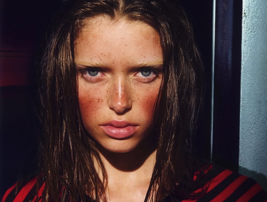 Close-up portrait of a woman with freckles and blue eyes, wearing a red and black striped shirt, with dark hair and soft lighting.