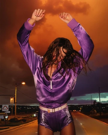 Woman in purple tracksuit dancing on a road at night with dramatic sky and street lights creating a dynamic fashion lifestyle image.