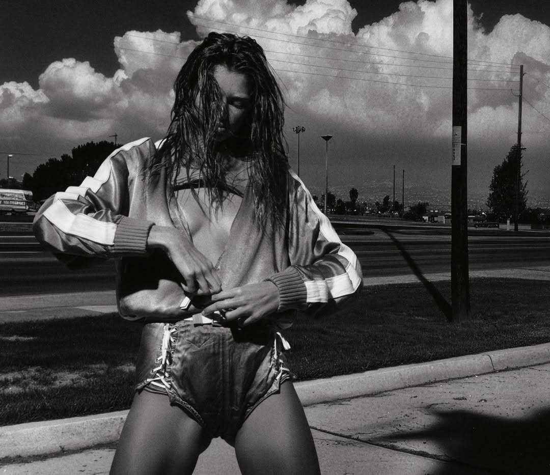 Black and white portrait of a long-haired adult woman wearing a striped jacket and shorts on a roadside with moody sky.