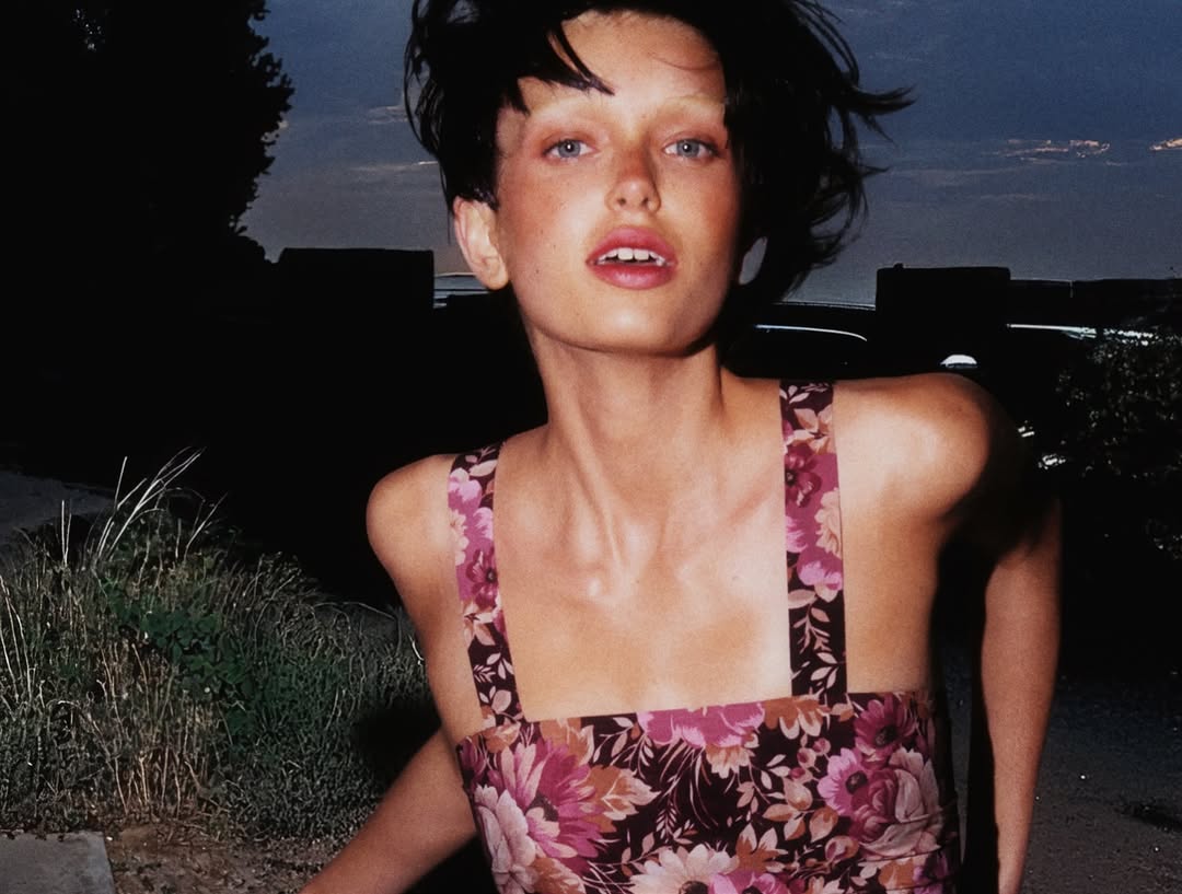 Portrait of a brunette woman in a floral dress outdoors at night with illuminated buildings in the distance, captured with a medium shot.