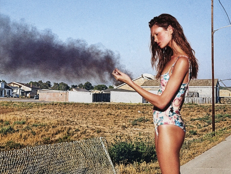 Woman in floral swimsuit standing outdoors by a road with smoke rising over buildings in the background.