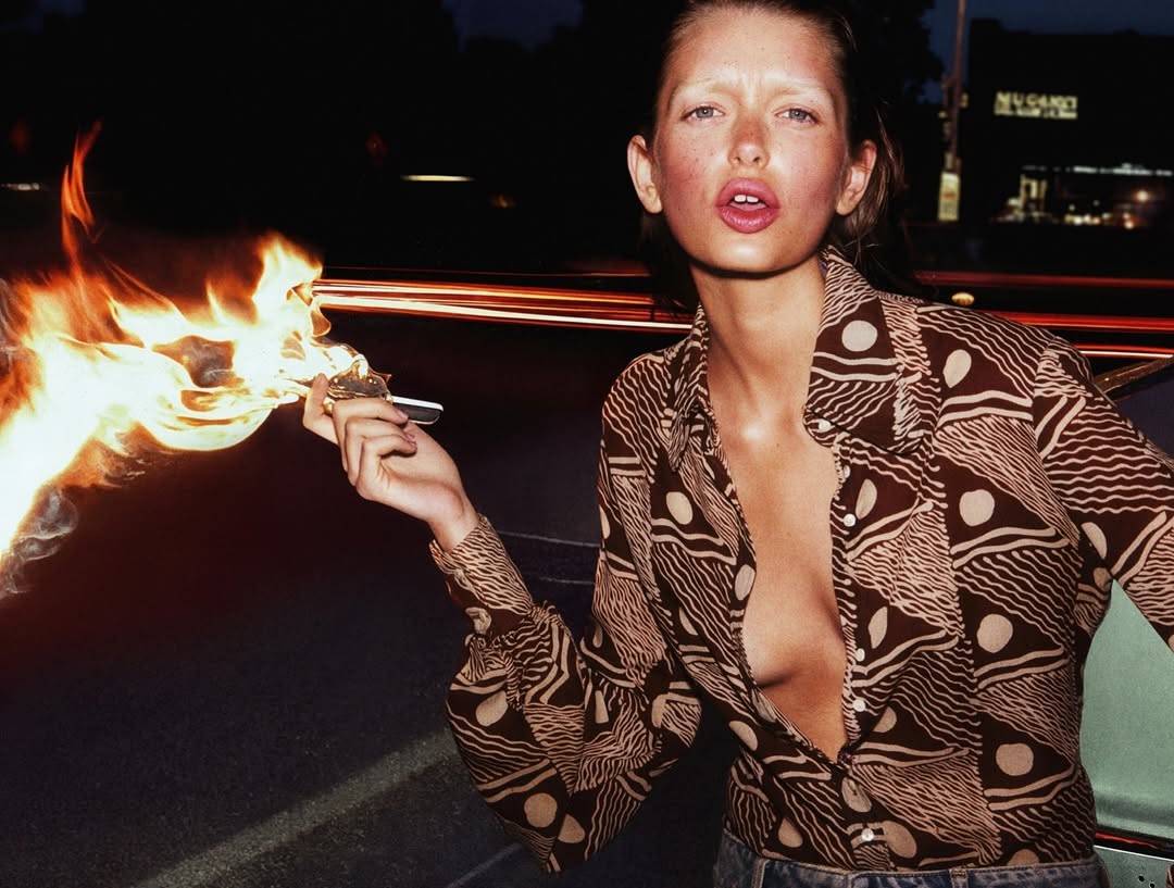 Adult female poses in outdoor night shot, holding burning cigarette with car in background, showcasing fashion and a lifestyle feel.