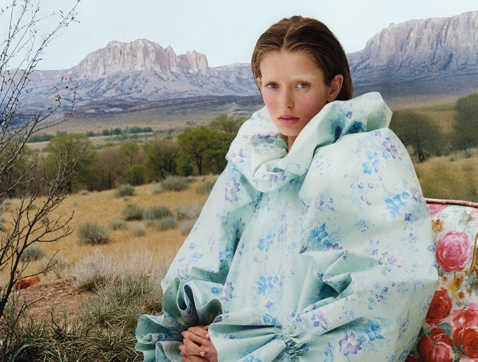 Female model in floral dress seated outdoors against a mountain backdrop. Fashion editorial shot with natural light and soft focus.