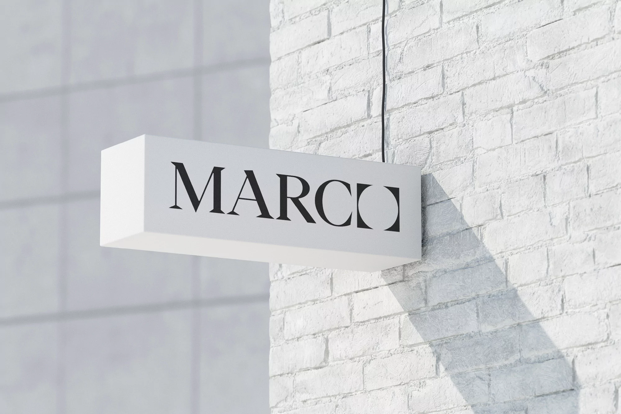 A rectangular white sign with the word 'MARCO' in bold black letters, hanging on a white brick wall with light shadows.