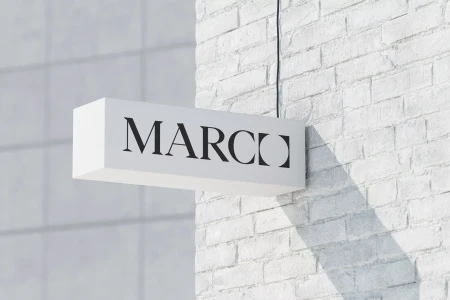 White rectangular projecting sign featuring the black brand name Marco mounted on a painted brick wall exterior.