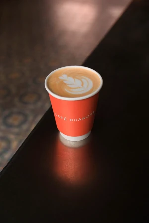 A single-use coffee cup with latte art sits on a dark surface with light reflecting off the table. The cup has the logo for Cafe Nuances.