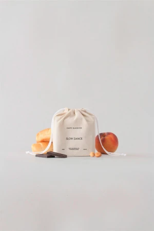 Still life photo featuring a cloth bag, a ripe peach, two hazelnuts, a piece of chocolate, and pastry rolls on a white background.