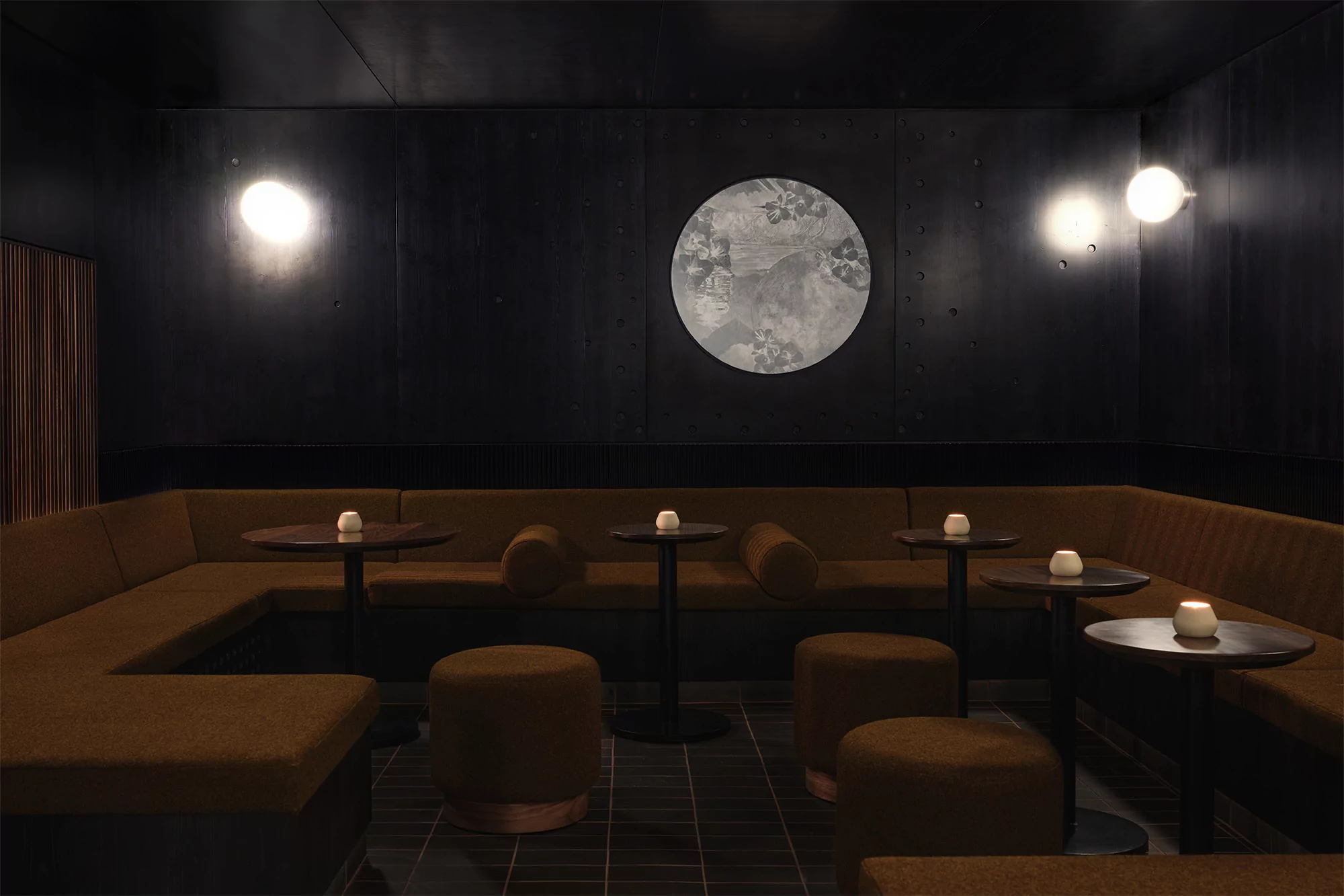 Stylish bar interior with dark walls, upholstered seating, small tables with candle lights, and a large round decorative wall piece.