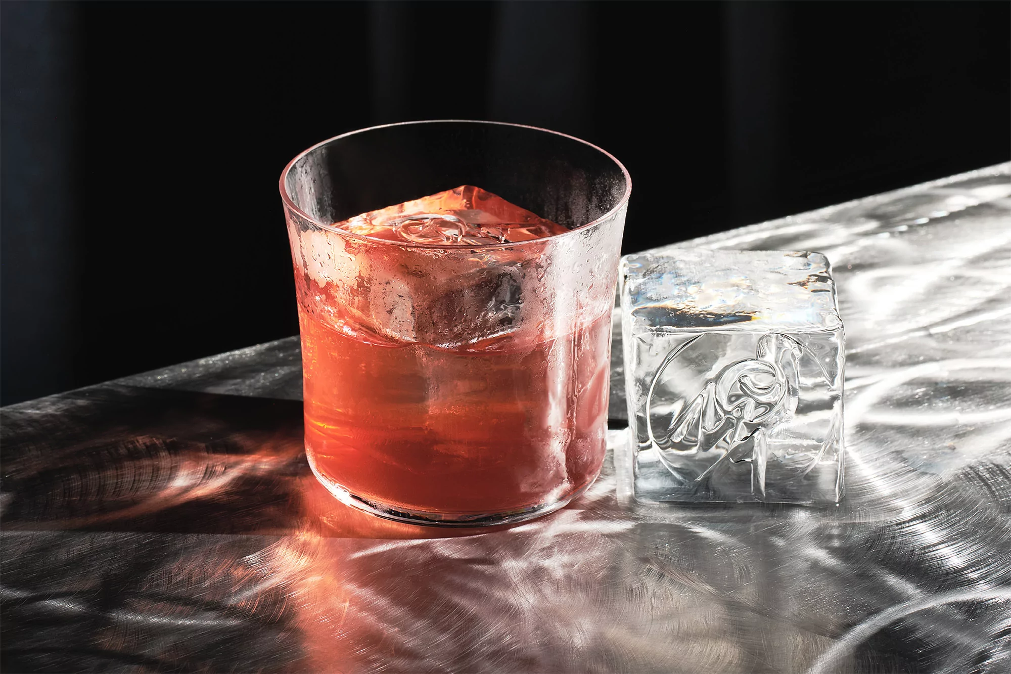 A glass of a reddish beverage with ice cubes sits on a metallic surface next to a large clear ice cube.