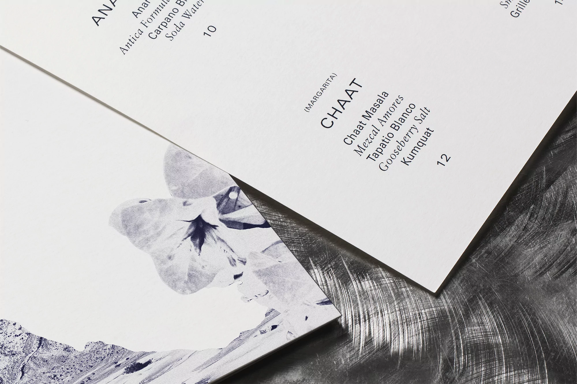 Overhead shot of cocktail menus on a brushed steel surface, featuring drink names and artistic mountain and flower illustrations.