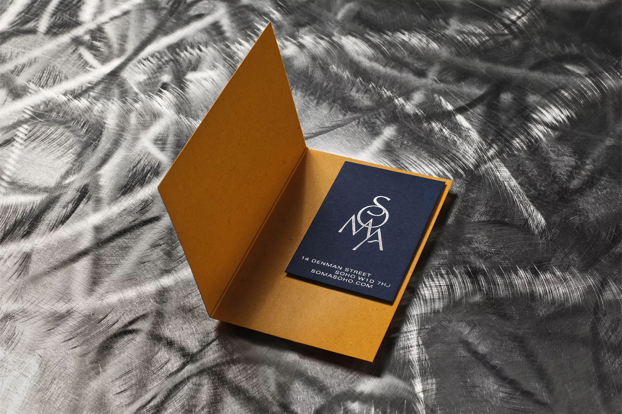 Studio shot of an open mustard-colored card revealing a small navy business card with minimalist white text and logo, set on a textured background.
