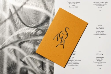 Orange card with SOMA logo resting on a printed white menu page over a textured metallic surface
