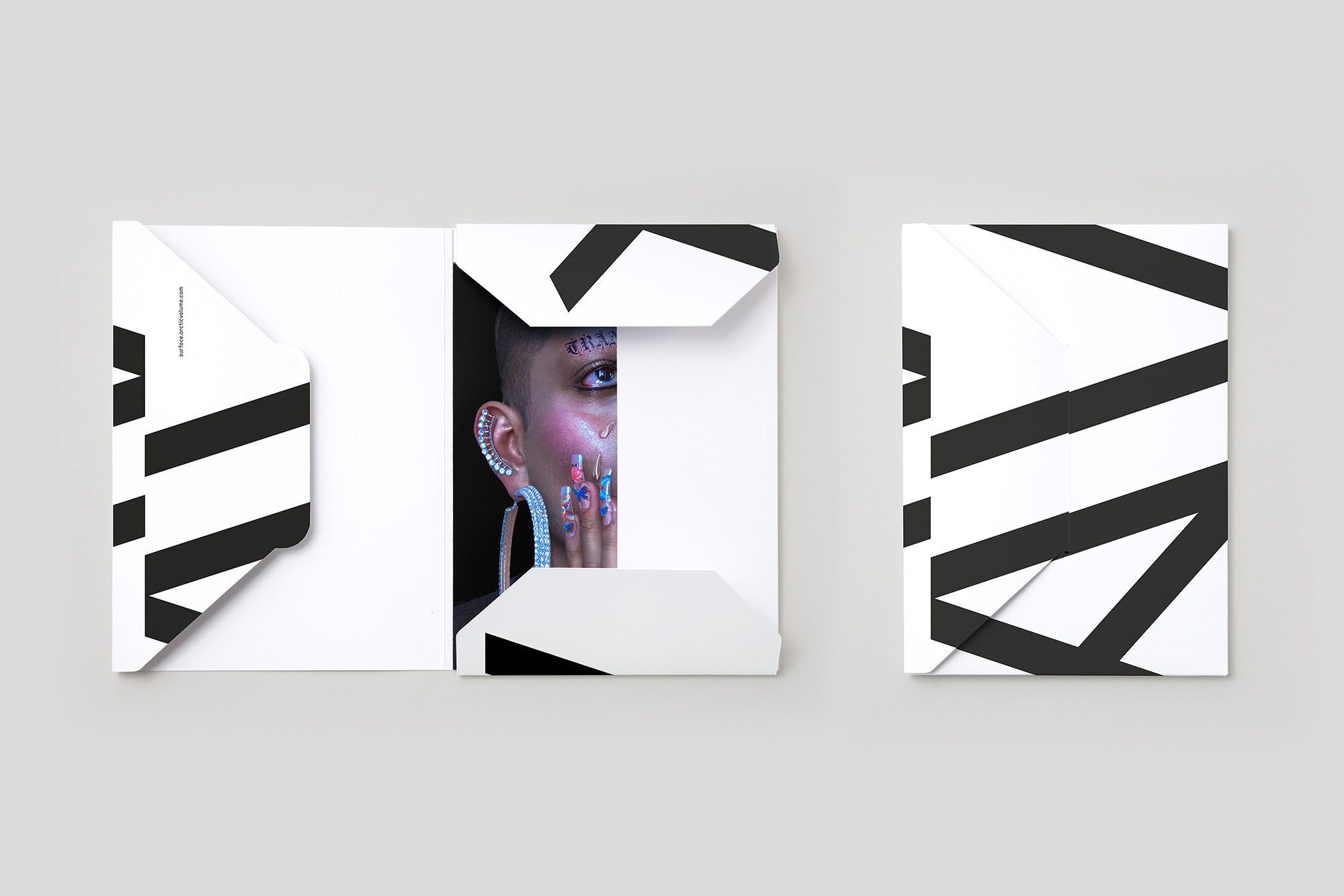 Close-up of person's face with makeup and jewelry partially visible through white paper cutout with black geometric design, on a gray background.