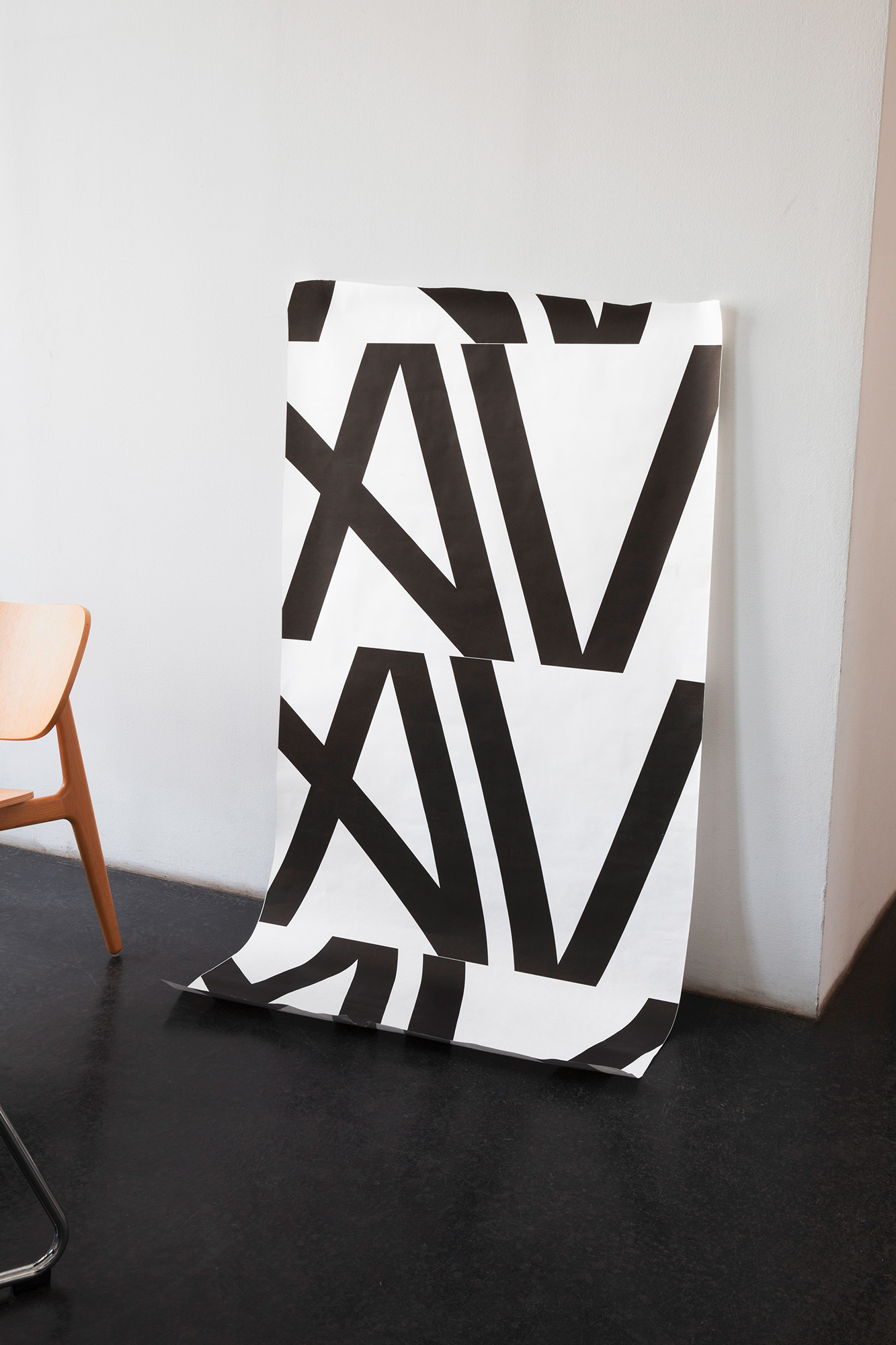 Roll of wallpaper with a graphic pattern leans against a white wall on a dark floor, next to a wooden chair.