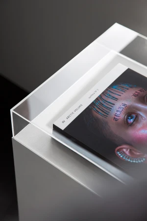 Fashion magazine with a person's face with body art on the cover, resting in a modern clear display stand with a dark background.