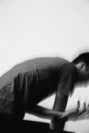 Monochrome photograph of an adult male leaning forward with hands clasped in front of a white wall, showcasing motion blur.