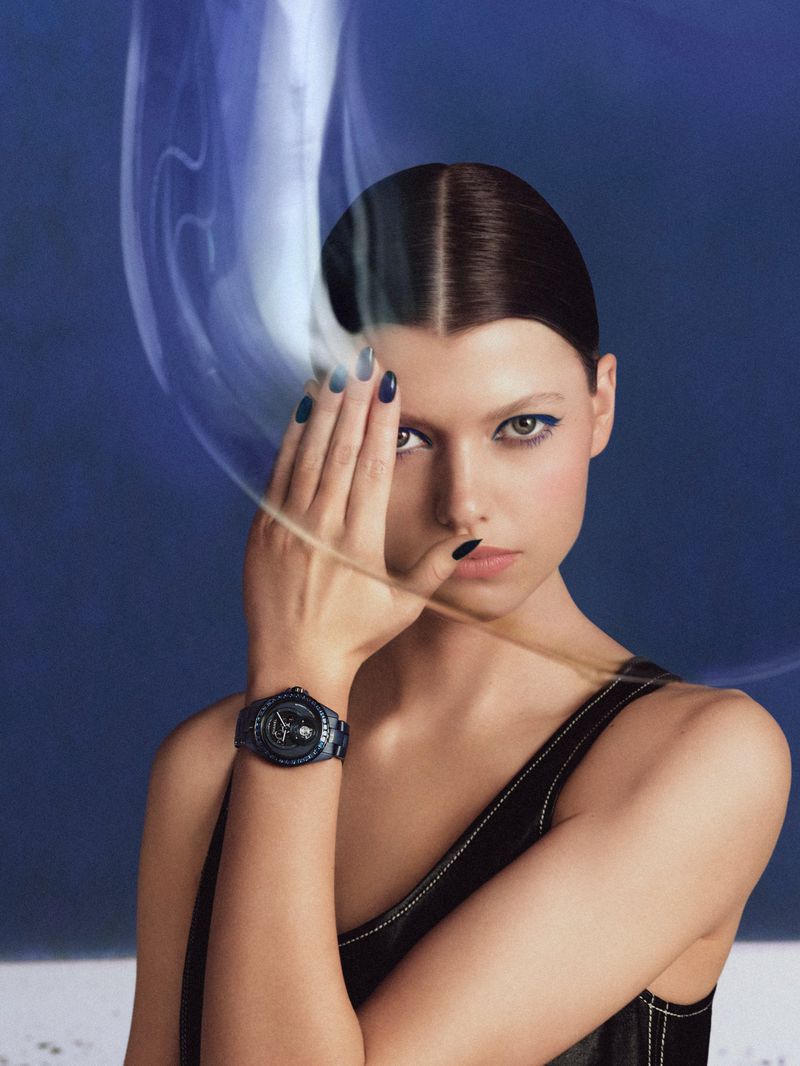 Close-up studio shot of a woman modeling a wristwatch. She is wearing a black top and blue eye shadow, with wispy blue trails around her.