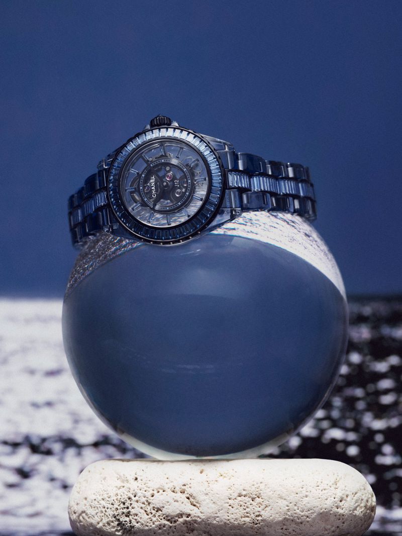 Elegant blue wristwatch displayed on a reflective orb atop a textured stone, set against a blurred, abstract background.