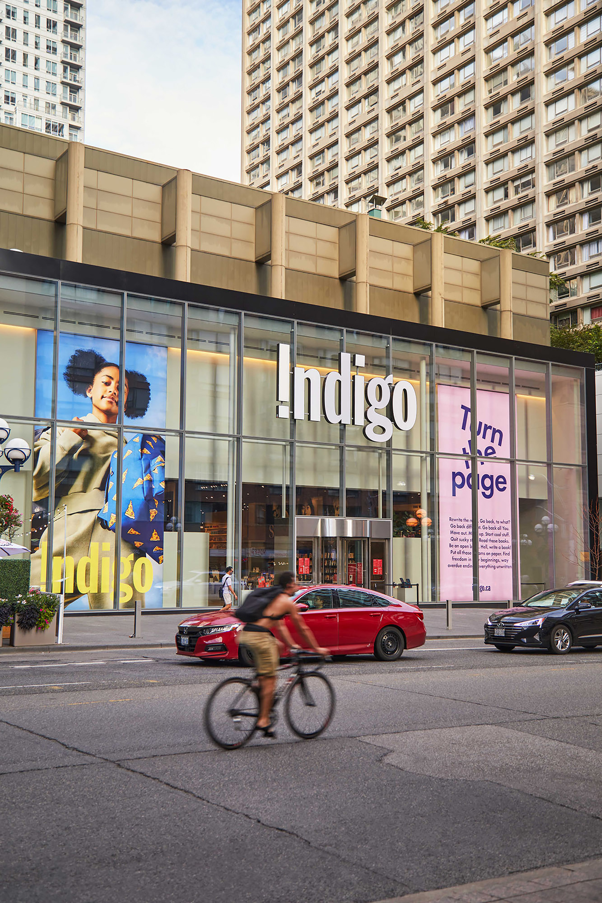 View of Indigo bookstore facade with people walking and cycling along the street, and vehicles passing by in Toronto, Canada.