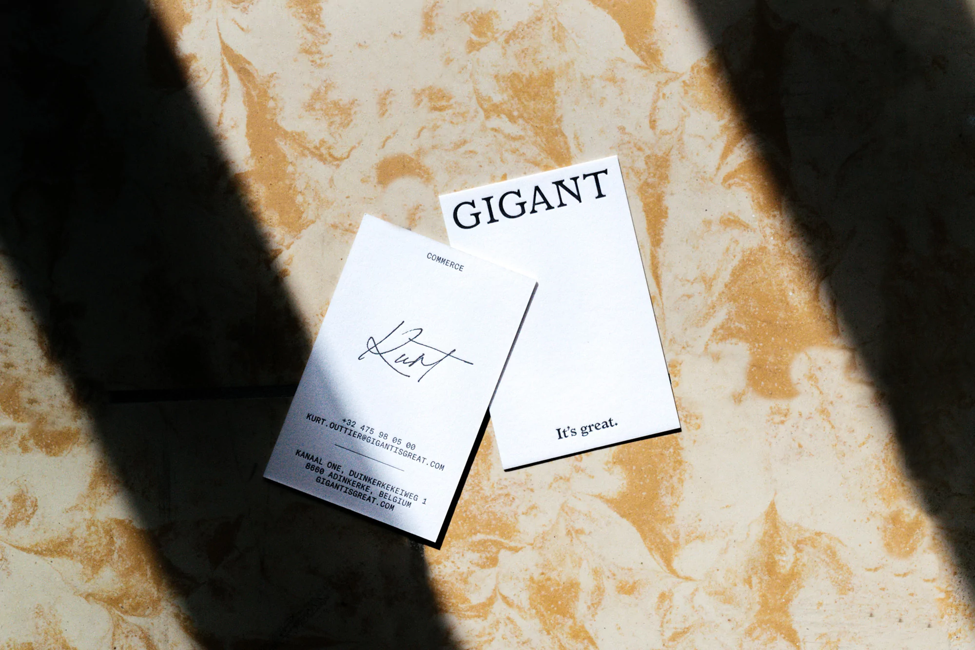 Two white business cards from Gigant Commerce, one with a signature, on a marble-like surface with shadows adding depth and visual interest.