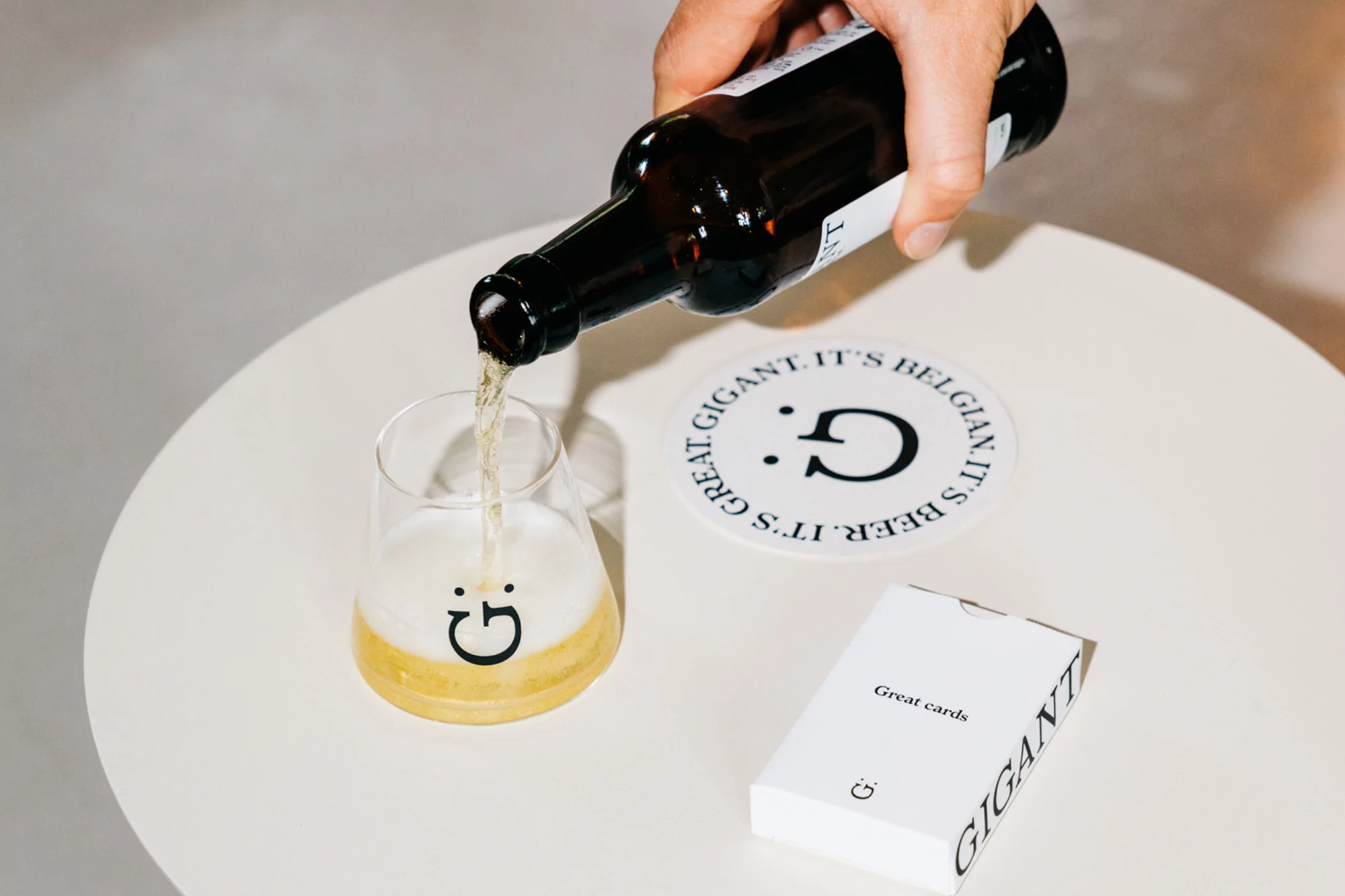 Beer bottle pouring into a branded glass with a coaster and card deck on a white table, capturing a moment of leisure and branding.