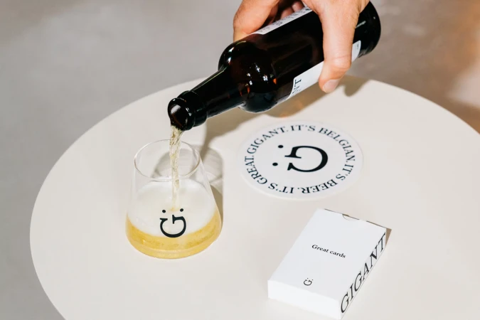 Beer bottle pouring into a branded glass with a coaster and card deck on a white table, capturing a moment of leisure and branding.