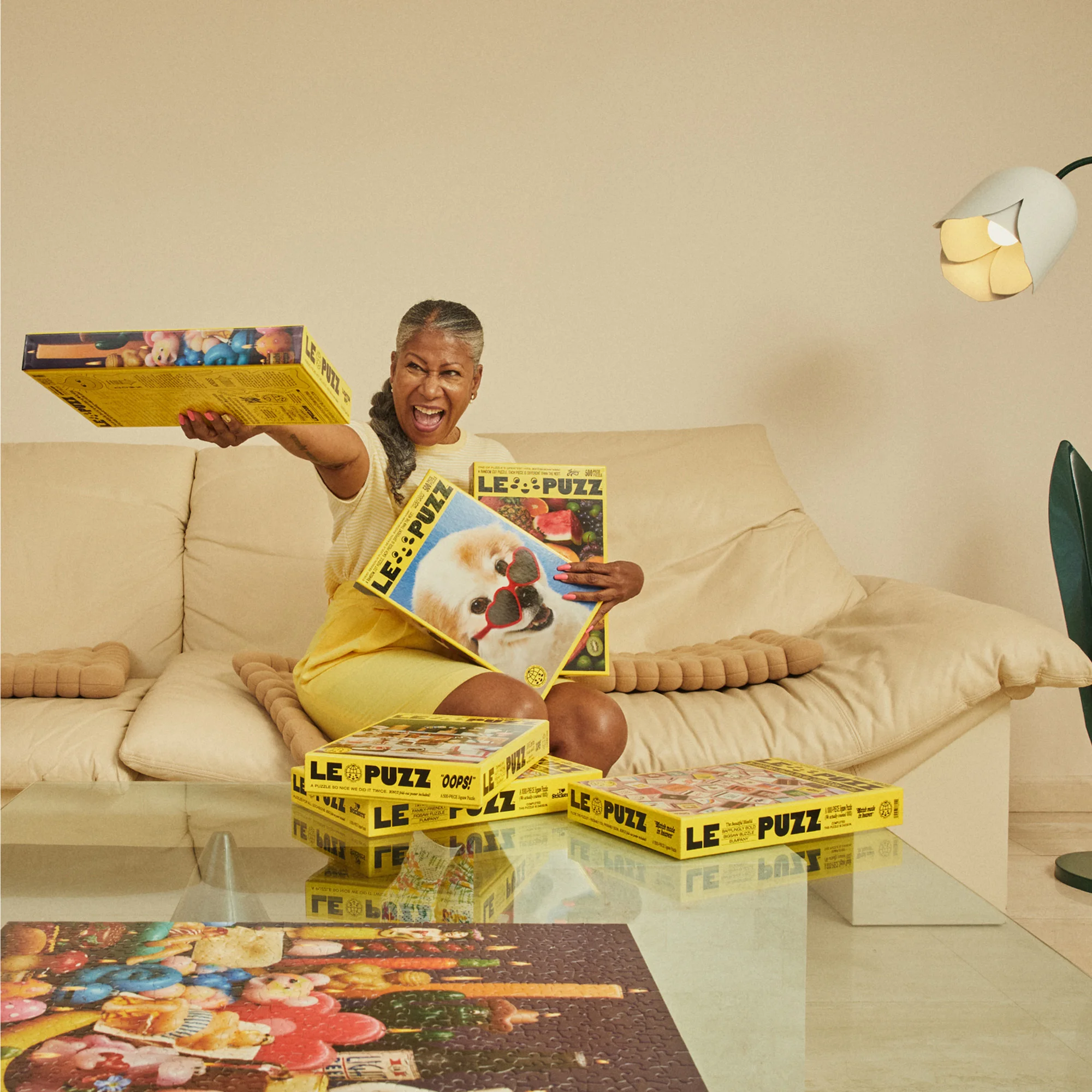 Excited mature woman in yellow dress poses with Le Puzz jigsaw puzzle boxes indoors, creating a fun and colorful lifestyle image.