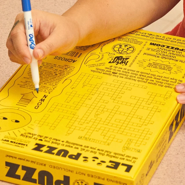 Hands holding blue marker over a yellow jigsaw puzzle box with crossword puzzle grid, alien illustration, and text, creating a playful scene.