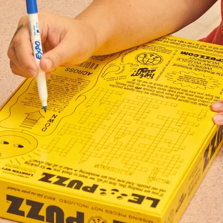 Hands holding blue marker over a yellow jigsaw puzzle box with crossword puzzle grid, alien illustration, and text, creating a playful scene.