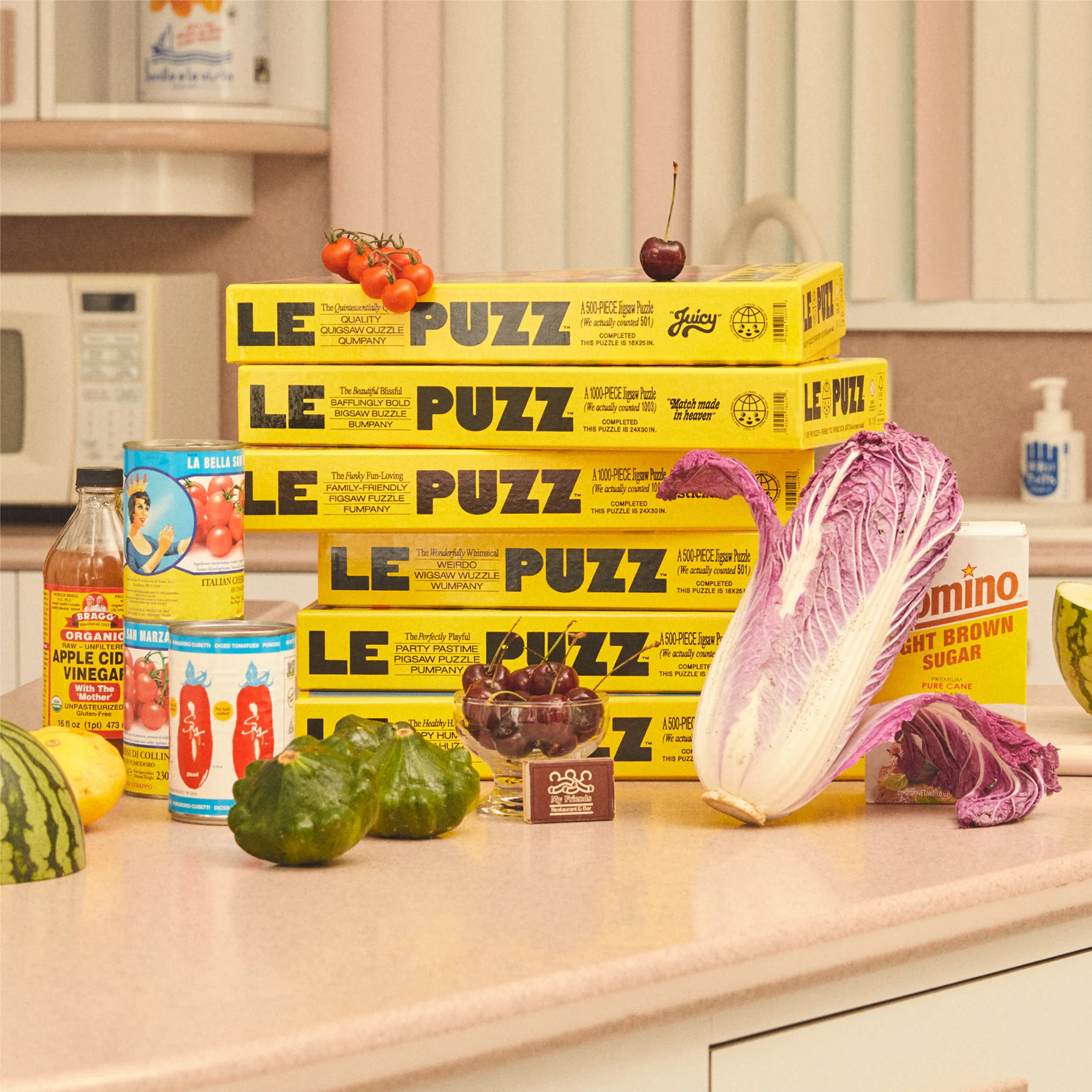 Still life of food and Le Puzz puzzle boxes, including cherries, tomatoes, lettuce, squash, and cooking ingredients on a pink countertop.