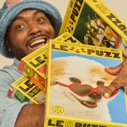 Smiling man in a blue denim bucket hat holding several colourful Le Puzz jigsaw puzzle boxes including one with a dog wearing heart glasses.