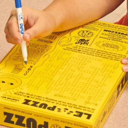 Person holding a blue dry-erase marker writing on a yellow Le Puzz jigsaw puzzle box featuring a printed crossword design.