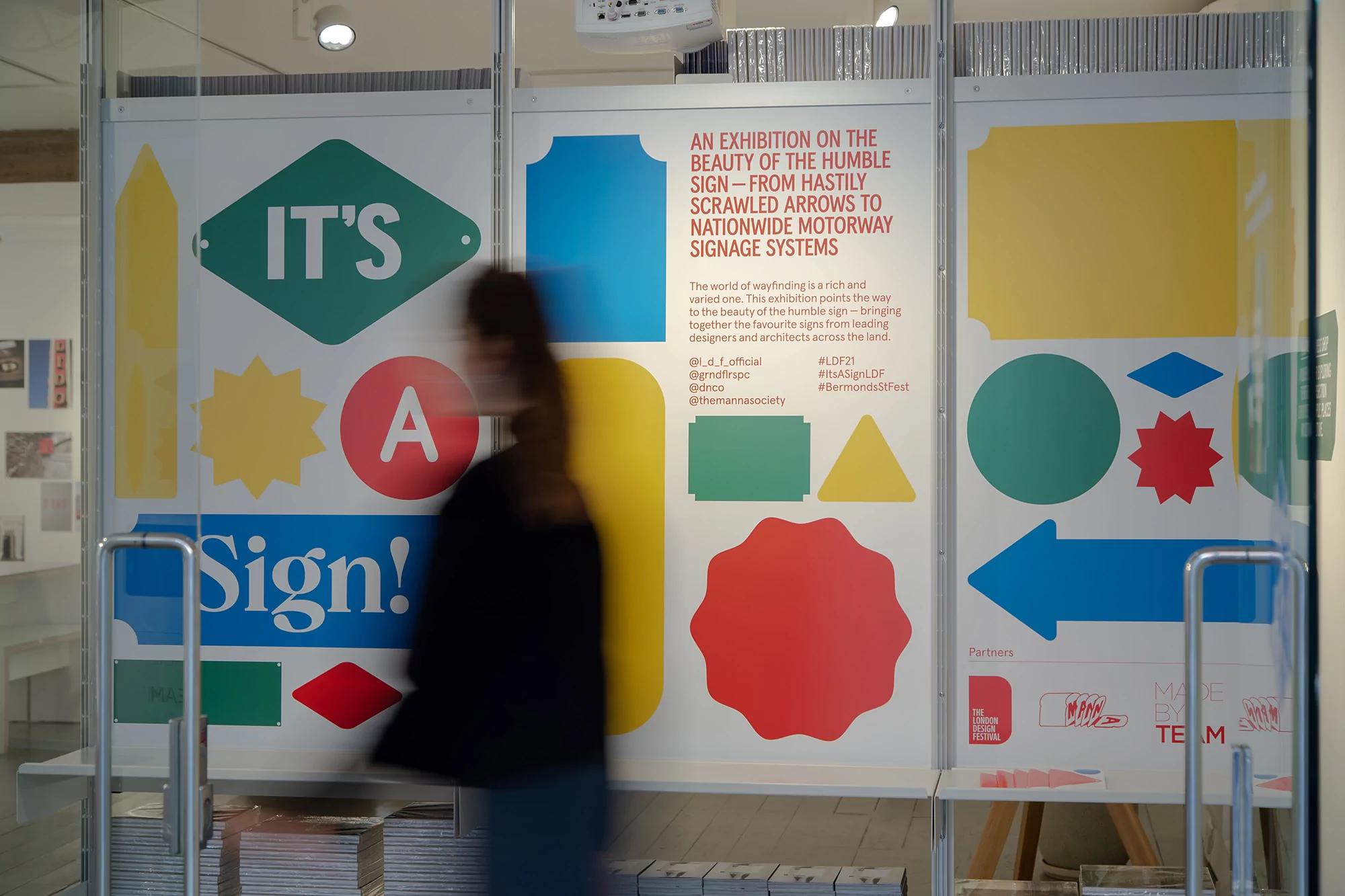 Exhibition display featuring colorful sign designs and typography, visible through the glass doors of a brightly lit interior space.