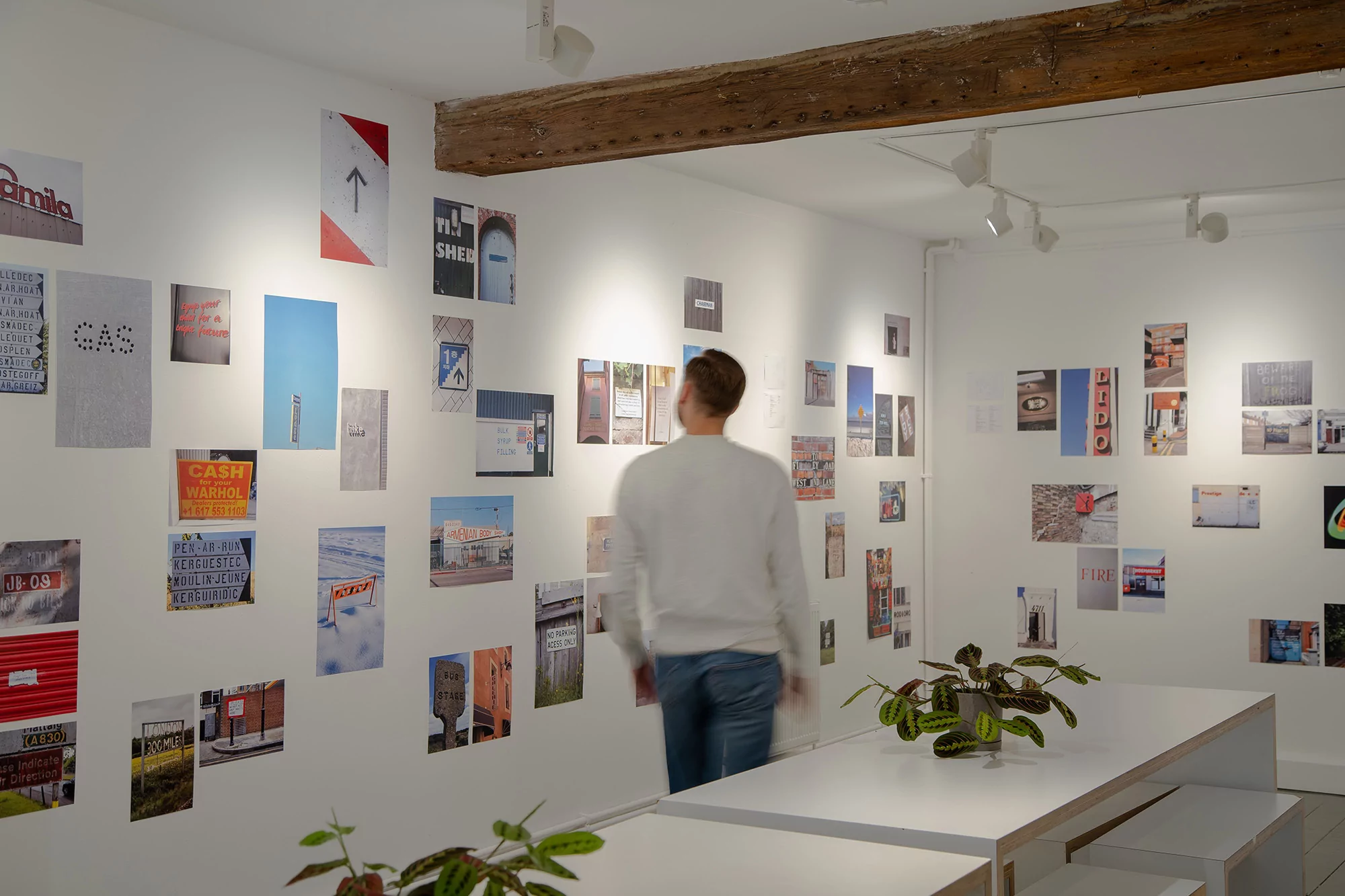 Man in white sweater looks at a wall of photographs in an art gallery with white walls, wooden beam, track lighting, and white tables.