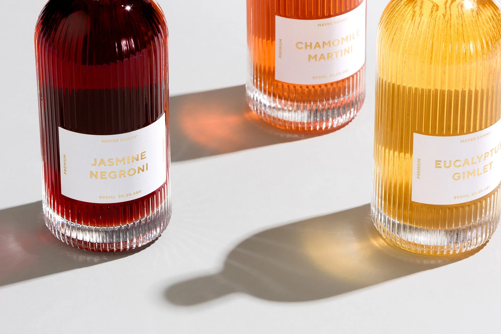 Three glass bottles of colorful cocktails, including Jasmine Negroni, Chamomile Martini, and Eucalyptus Gimlet, are on a white surface.