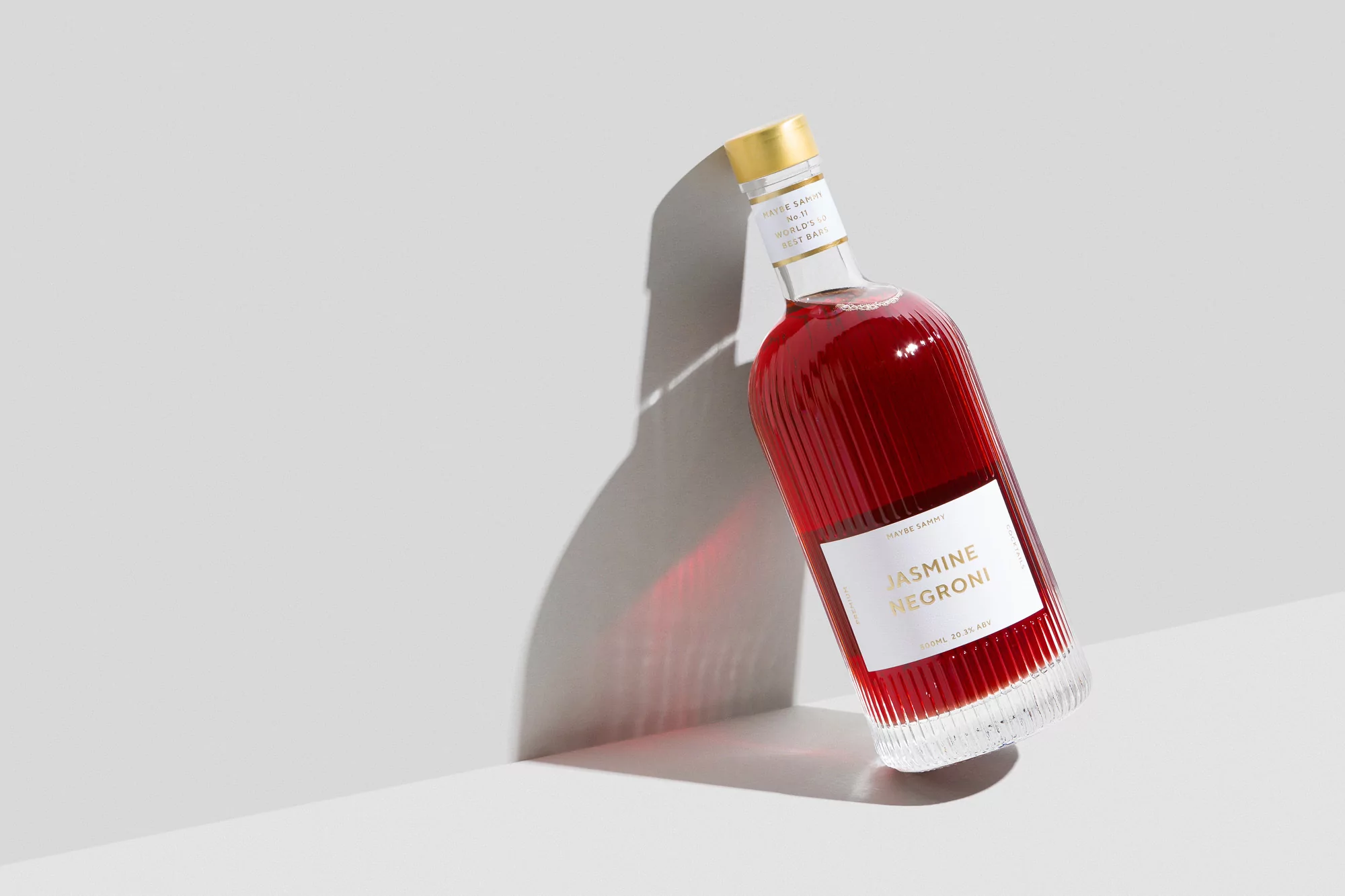 Studio shot of a tilted glass bottle filled with red liquid and a gold cap, casting a long shadow against a neutral background.